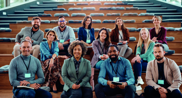 A group of professionals sitting on wooden steps in an auditorium, smiling at the camera.
